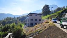 3-story modern residential building on a hillside, surrounded by trees and mountains. The building has a raised ground floor and is under construction, with construction equipment and materials visible on the site.