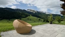 The image shows an outdoor patio area with a wicker chair overlooking a scenic mountain landscape. The patio is made of stone tiles and there is a small potted plant nearby. In the background, there are snow-capped mountains and a forested valley with gre