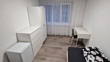 Spacious bedroom with white furniture, including a dresser, desk, and chair. The room has hardwood flooring and large windows with sheer curtains, providing natural light. A small refrigerator and a washing machine are also visible in the room.