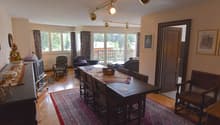 The image shows a spacious open-plan living and dining area. The room has large windows that provide a view of a balcony or terrace overlooking a wooded area. The living room features comfortable sofas and chairs, while the dining area has a wooden table