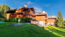 2-story wooden chalet-style house with balconies, surrounded by lush green lawn and trees, set against a blue sky