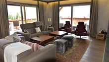 Spacious living room with large windows, hardwood floors, comfortable sofas and armchairs, wooden coffee table, and a balcony or terrace visible through the windows.