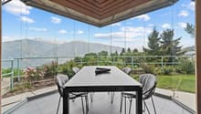 Spacious dining area with a large glass-enclosed patio, offering a panoramic view of the surrounding mountains and lush greenery. The room features a modern black dining table and chairs, with a wooden ceiling and beams providing a warm, rustic ambiance.
