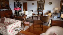 Spacious living room with a mix of traditional and vintage furniture, including a floral patterned sofa, wooden chairs, a coffee table, and a bookshelf. The room features hardwood floors, white walls, and decorative elements such as a large mirror, artwor