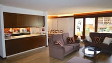 modern kitchen with wooden cabinets, dining area with chairs, two couches, coffee table with books, lamp, large windows