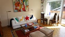 living room: white couch with colorful pillows, glass coffee table, wooden floor, abstract painting on wall, large window, balcony with wooden railing, view of mountains