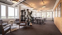 Modern office room with open layout, carpeted floor, gray chairs, tables, glass windows, pendant lighting, and greenery