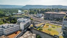 Aerial view of a city with a mix of modern and older buildings, surrounded by hills and greenery. The image shows a train station, parking areas, and green roofs on some of the buildings.