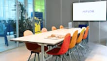 Empty meeting room with glass walls, long white table, orange and blue chairs, screen, whiteboard