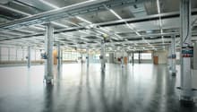 Empty warehouse with metal beams, exposed pipes, large windows, concrete floor.