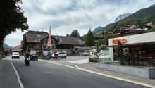 A street with a motorcycle and cars passing by, a building with a flag, a mountain in the background, and a shop with the name 'COOP'.