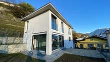 Modern white house, large glass windows, modern architecture, sloping ground with vegetation, small porch with glass doors, concrete fence, mountains in the background.