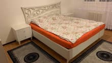 a bed with white headboard, white cabinet, and two rugs, white wall, window with curtains