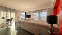 Modern and spacious bedroom with a king-size bed, large windows with white curtains, two armchairs, side table with lamp, small table with chairs, and a TV mounted on the wall. A rug is placed near the chairs.