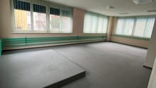 An empty room with gray carpet floor, white ceiling, large windows with metal blinds, and an elevated platform on the left.