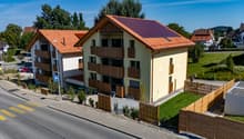 Two-storey house, red roof, yellow exterior, solar panels, wooden balconies, gated entrance, green lawn, road with pedestrian crossing