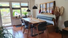 wooden dining table with 6 chairs, black bench with cushions, white wall, big windows, floor heating, plants