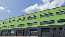 A multi-story commercial building with a green exterior and large windows. The building has multiple garage doors on the ground floor, indicating it may be used for industrial or warehouse purposes.