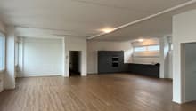 an empty, spacious room with wooden floors, white walls, and a kitchen area in the back