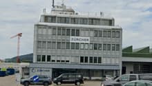 Multi-story office building with the Zurcher logo prominently displayed, surrounded by parked cars in a parking lot.