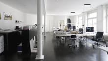 Large open office space with multiple desks, computers, and office equipment. The room has high ceilings, white walls, and dark hardwood floors. There is a dishwasher visible in the corner of the room.