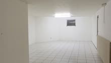 The image shows a large, empty room with white walls and a tiled floor. The room appears to be in a commercial or industrial building, with a simple light fixture visible on the ceiling.