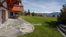 2-story wooden chalet-style house with a large balcony, surrounded by a well-maintained lawn and landscaping. The house has a stone patio in the foreground and offers a scenic view of snow-capped mountains in the background.