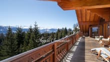 Wooden balcony with a view of snow-capped mountains and a forested landscape. The balcony has a railing with a decorative pattern and there are wooden chairs and furniture on the balcony.