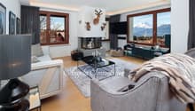 fireplace, 3 deer heads mounted on wall, 3 windows, 2 couches, gray area rug, glass table, gray armchair with fur blanket