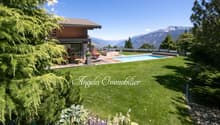 Luxury house with wooden exterior, surrounded by lush greenery and mountains in the background. The property features a swimming pool and offers a scenic view of the surrounding landscape.