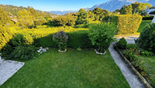well-maintained garden with green lawn, hedges, and a gravel path, surrounded by trees and bushes, with a distant view of mountains
