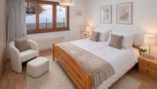 Bedroom with wooden bed, white bedding, wooden nightstands, white chair, white ottoman, white rug, white walls, large window with curtains, radiator, lamps, framed pictures, flowers