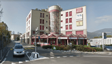 A multi-story building with a modern architectural design, featuring glass windows and red shutters. The building has a signboard with the text 'Il Caffe' and other advertisements. There are cars driving on the road in front of the building, and a pole wi