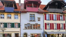 The image shows a row of traditional, colorful buildings with red tile roofs, wooden shutters, and decorative architectural elements. The buildings appear to be located in a town or city setting, with a cobblestone street in the foreground. The buildings