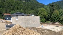 The image shows a house under construction in a mountainous, forested area. The foundation and walls of the house are visible, and there appears to be a garage or other structure next to the main building. The surrounding landscape is lush and green, with