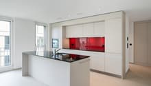 Modern, open-concept kitchen with white cabinets, red glass backsplash, black countertop, and built-in appliances including a dishwasher. Large windows provide natural light and access to a balcony or terrace.