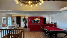 The image shows a large, open-concept living space with a dining area, kitchen, and entertainment area. The space features a high, vaulted ceiling with a large, ornate chandelier. The walls are a mix of white and red, with wooden beams and accents. The ki