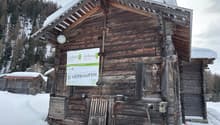 Rustic wooden cabin with a sloped roof, surrounded by snow-covered trees. The cabin has a sign indicating it is for sale.