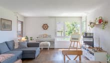 Spacious living room with large windows, grey sofa, wooden furniture, artwork, and potted plants. The room has a cozy and modern feel with a balcony or terrace visible through the windows.