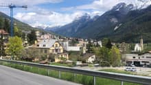 Mountain town with houses, mountains, road, cars, crane, trees, grass, sky