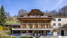 A traditional Swiss-style wooden building with a large balcony and terrace, housing a Gasthaus (inn or guesthouse) called 'Gasthaus St. Anton'. The building has a prominent sign displaying the name and is surrounded by trees and a grassy area.