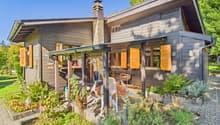 wood house, orange shutters, garden with plants, brick pathway, chimney, porch with table and chairs, grill, trees in the background