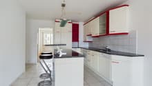 Spacious kitchen with white cabinets, red accents, granite countertops, tiled flooring, and a dishwasher. The kitchen is open to a dining area with a bar counter and stools.