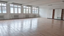 Empty room with large windows, wooden floor, white walls, built-in cabinets, radiators, fluorescent ceiling lights.