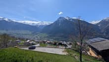 The image shows a scenic mountain landscape with snow-capped peaks, a small village nestled in the valley below, and a clear blue sky with a few clouds. There are solar panels visible on some of the buildings, indicating the use of renewable energy. The f