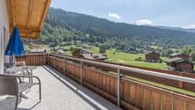 Wooden balcony with a view of a mountainous, forested landscape. The balcony has a table and chairs, and overlooks a village nestled in the valley below.