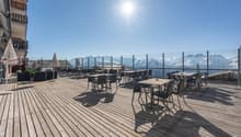 Outdoor dining area with wooden deck, tables and chairs, surrounded by glass railings offering a panoramic view of snow-capped mountains under a bright, sunny sky