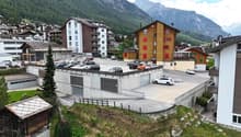 The image shows a residential area in a mountainous region. There are several multi-story apartment buildings and houses, some with colorful facades. There is a parking lot in the foreground with several cars parked. The surrounding landscape includes for