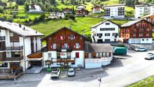 Mountain village complex with multiple buildings, wooden facades, and red shutters. One building has a restaurant sign. The complex is surrounded by a green hillside. Cars parked in front of the buildings.