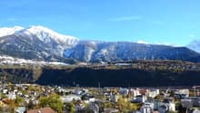 View of mountains with snow in background, surrounded by a town with various buildings, trees and greenery